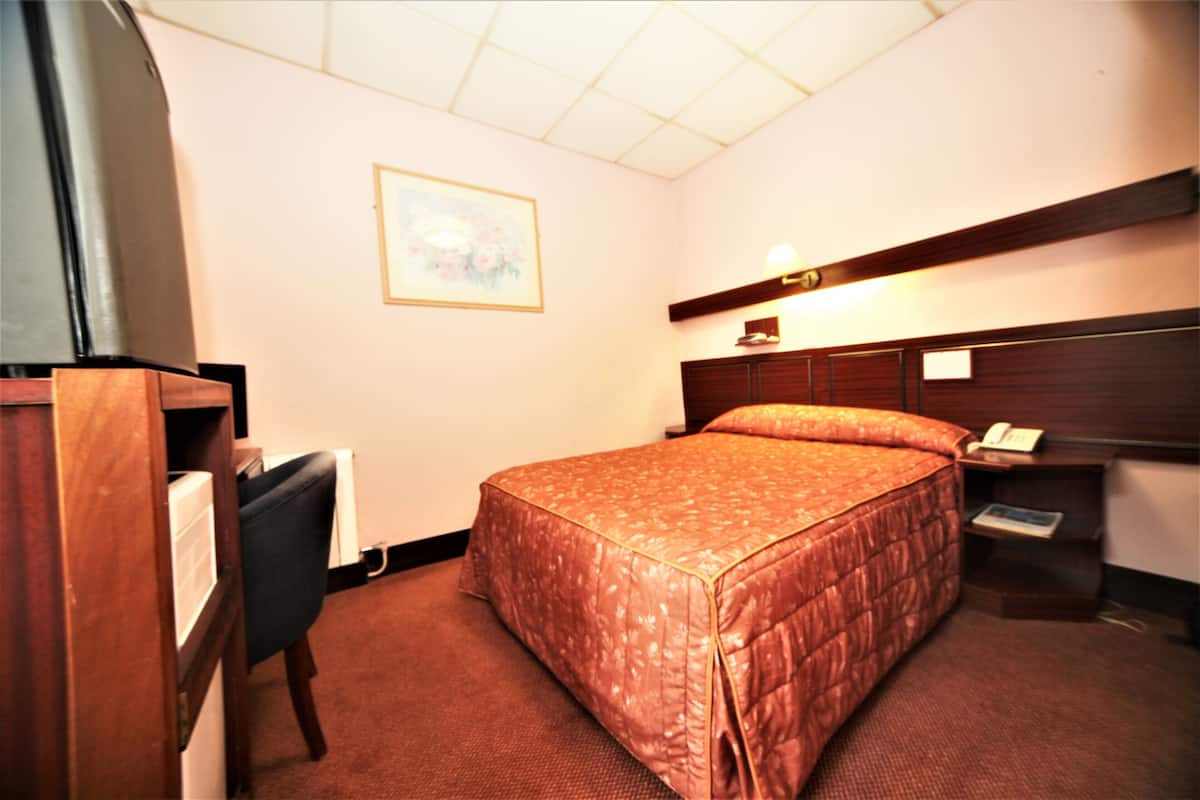 A hotel room with a bed, a desk, a chair, a nightstand, and a wall-mounted lamp.