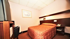 Habitación de hotel con cama, escritorio, silla, mesita de noche y lámpara de pared.