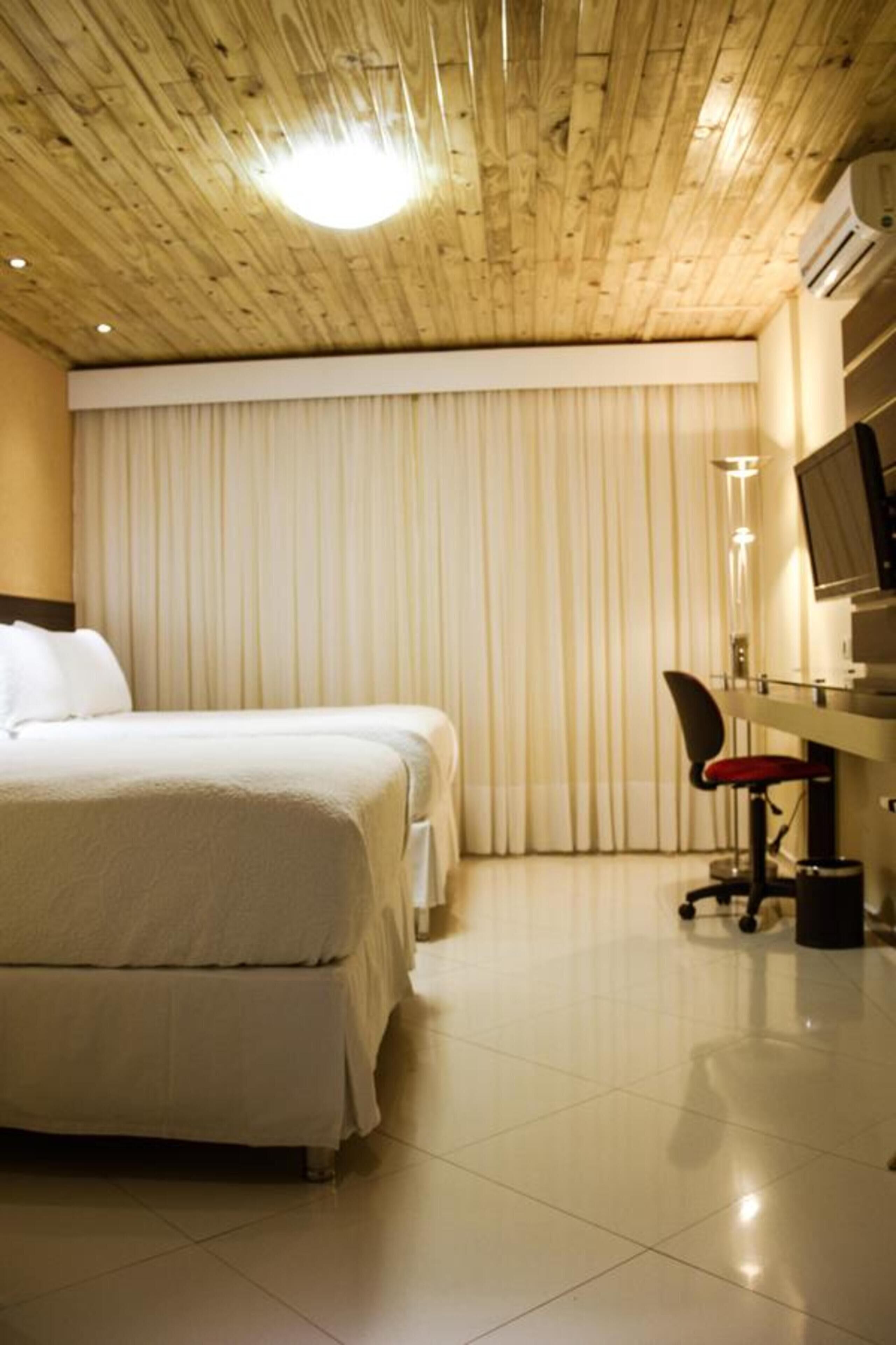 A hotel room with a wooden ceiling, a bed, a desk with a chair, and a large window with curtains.