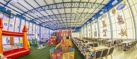 Un amplio espacio interior para juegos con techo alto, estructuras de recreación y mesas y sillas dispuestas para comer.