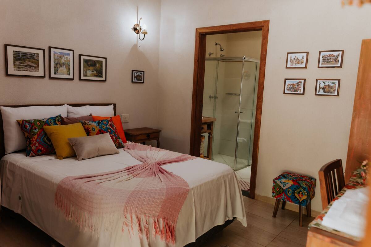 Un dormitorio con una cama, una mesita, una silla, cabina de ducha de vidrio y varias fotos enmarcadas en la pared.