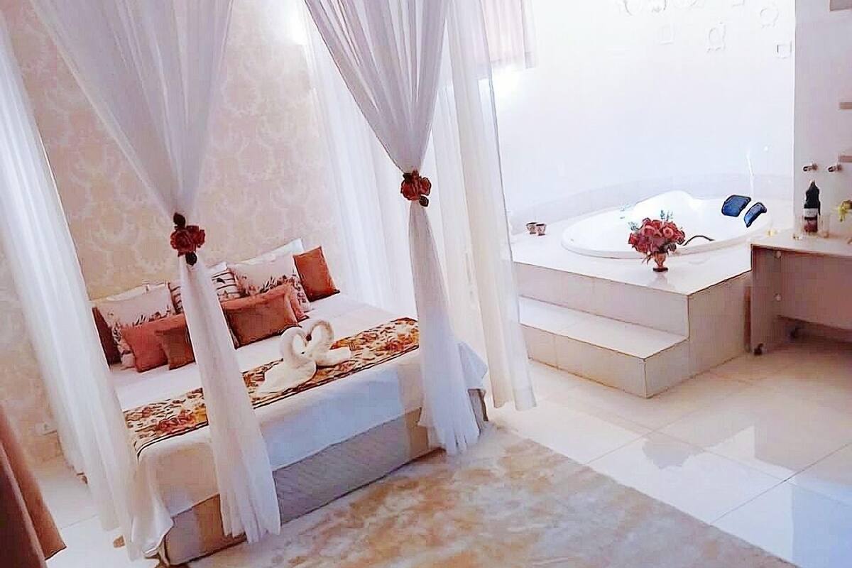 Un dormitorio moderno con cama con dosel, una zona de estar con una silla y un baño separado con bañera.
