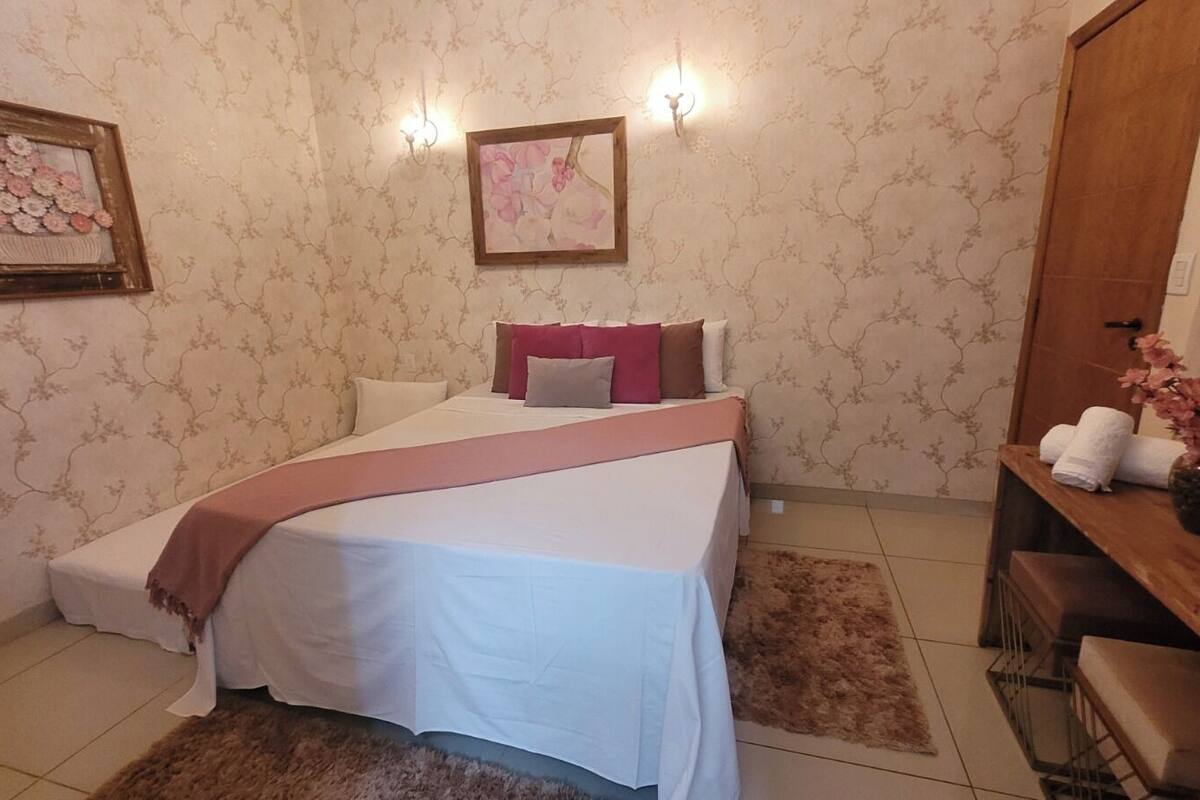 Un dormitorio ordenado con cama, almohadas, una mesita de noche y un buró.