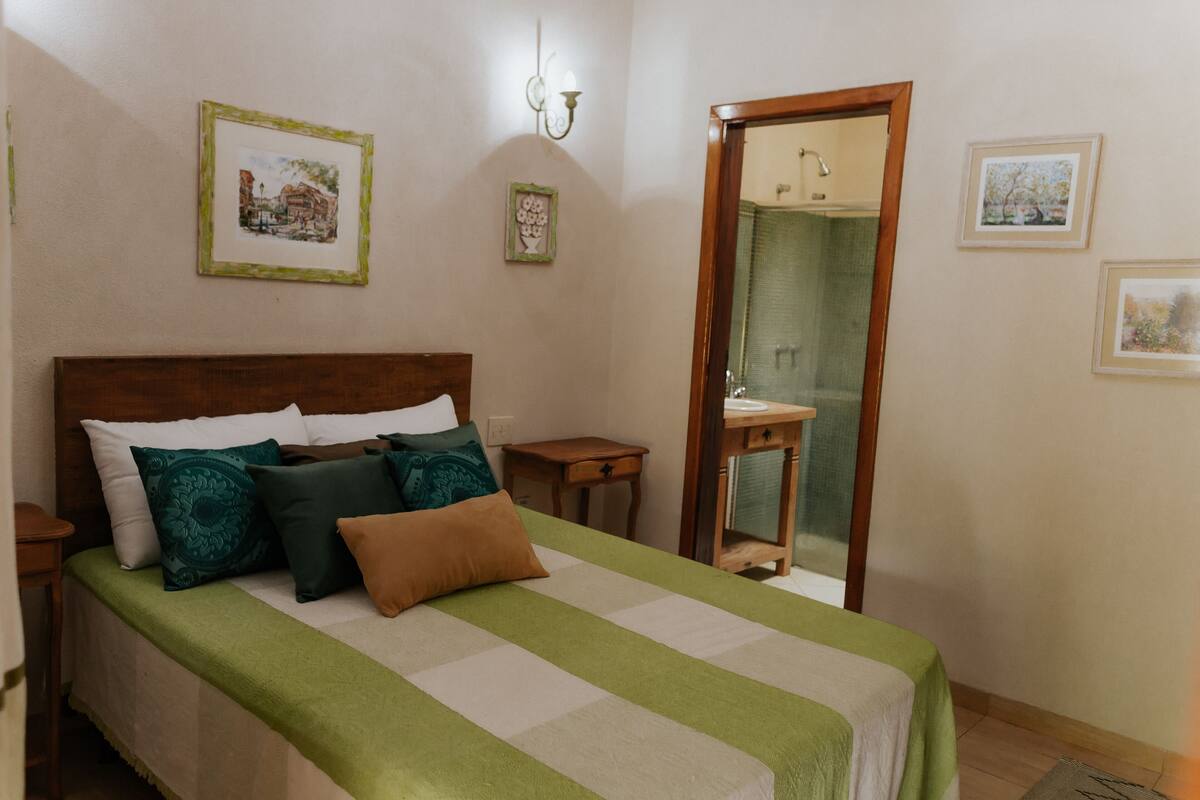 Una cama bien hecha con una colcha verde y blanca a rayas y varias almohadas. Se aprecia un cabecero de madera, una mesita de noche pequeña y un espejo en la pared.