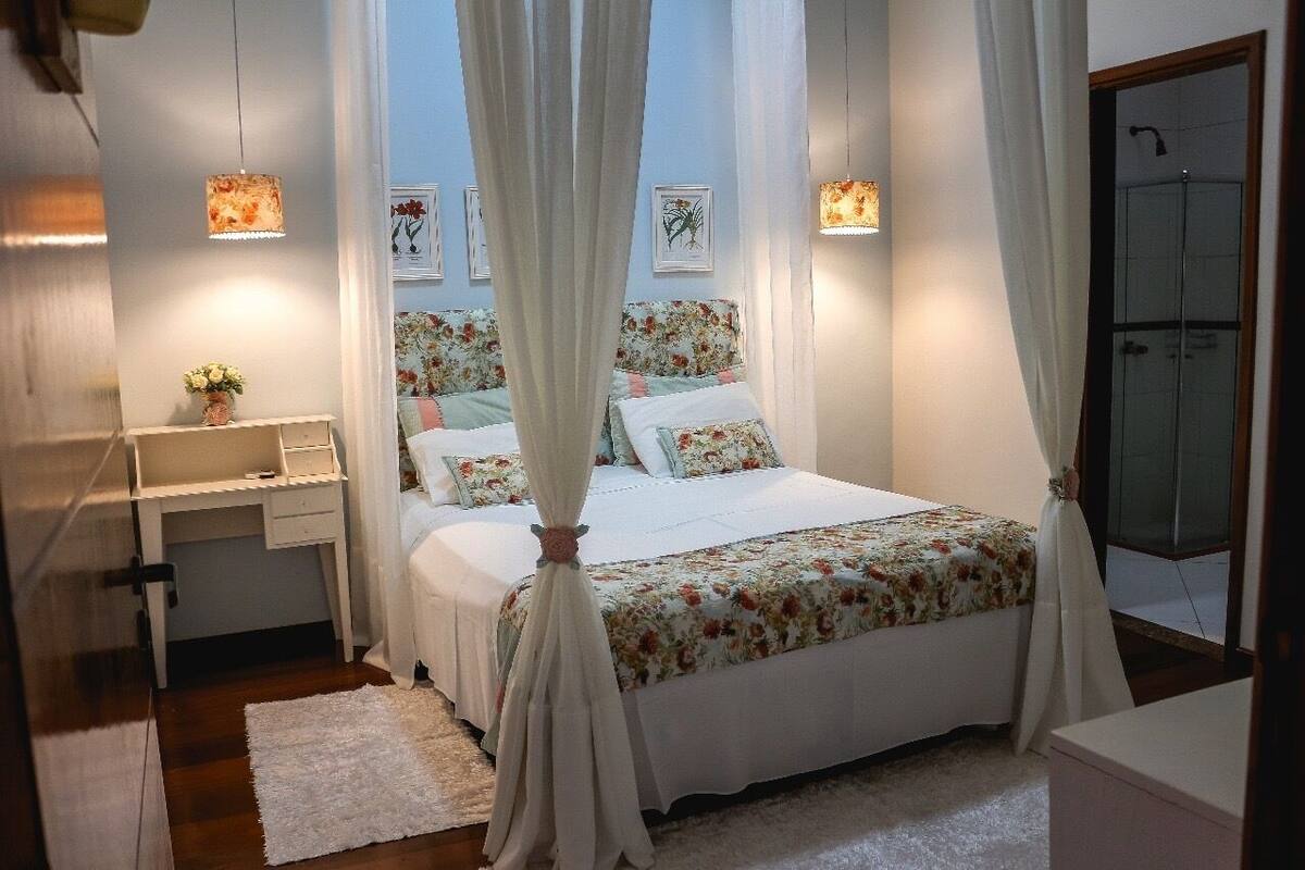 Un dormitorio con cama, mesita de noche y tocador. Hay una ventana con cortinas, una lámpara de pared y una puerta que da a un baño.
