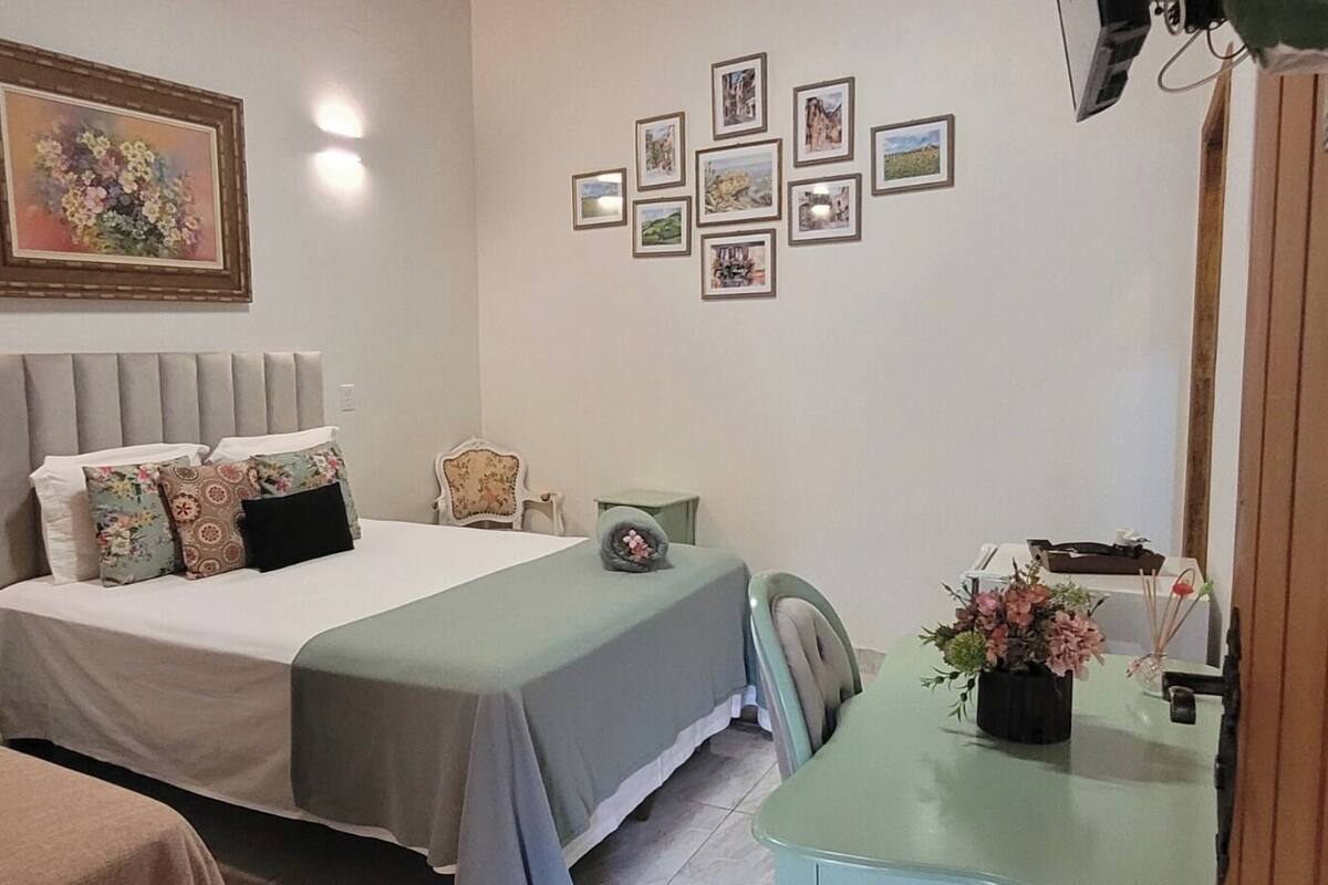 Un dormitorio con cama, mesitas de noche, una silla, un escritorio y una pared con cuadros.