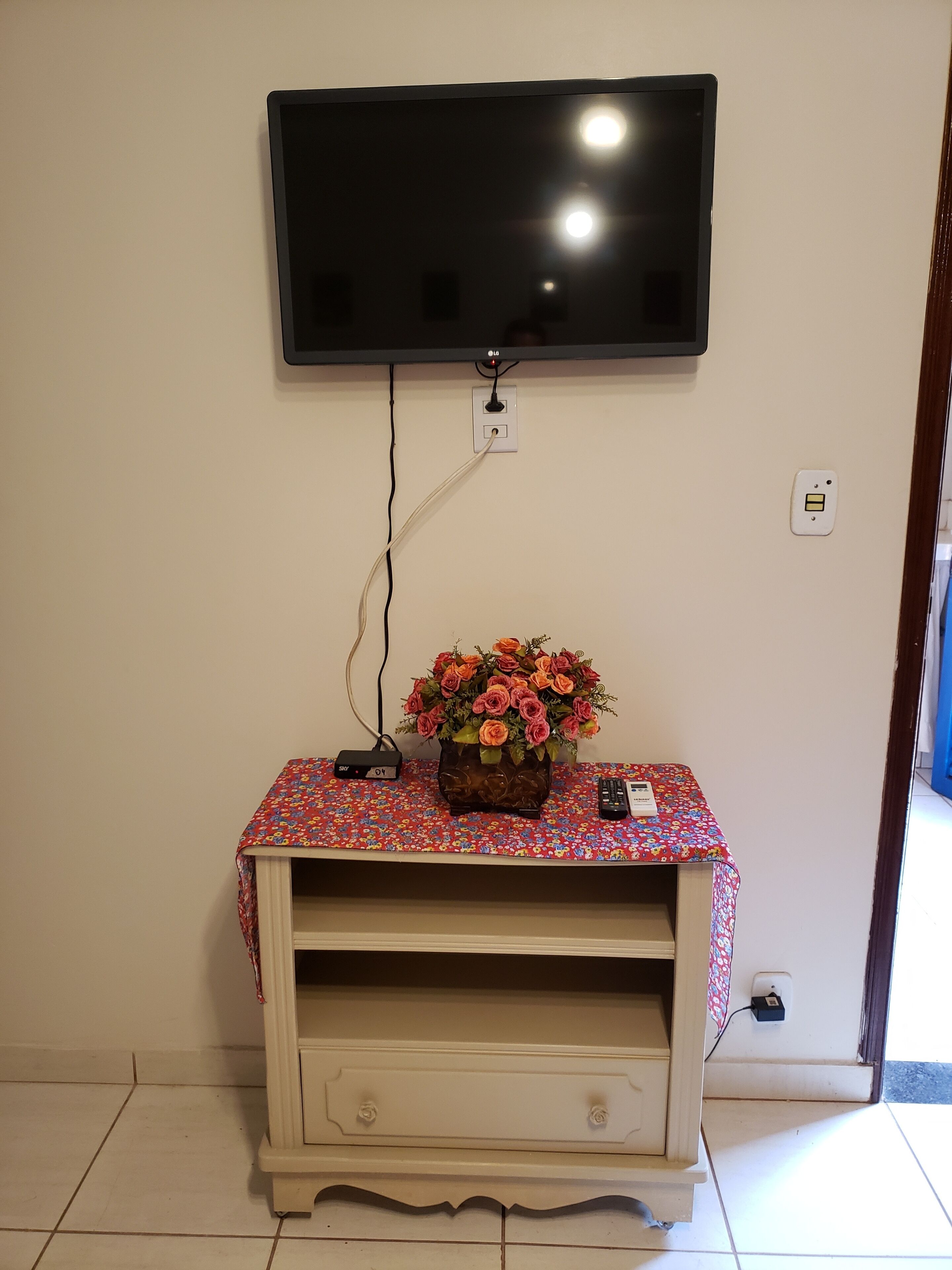 A television mounted on a wall above a wooden entertainment unit with a floral tablecloth.