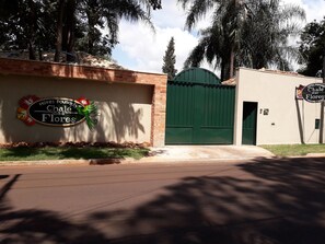 A hotel entrance with a green gate, a sign reading "Hotel Pousada Chale das Flores", and a brick wall with a decorative sign. - Hotel Pousada Chalé Das Flores (Sertaozinho)