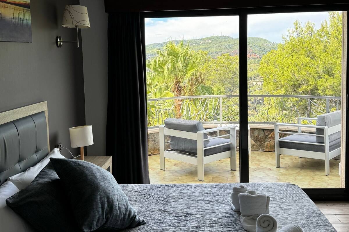 Een slaapkamer met een groot bed, een nachtkastje met een lamp, uitzicht op een balkon met stoelen, en een schilderij aan de muur.