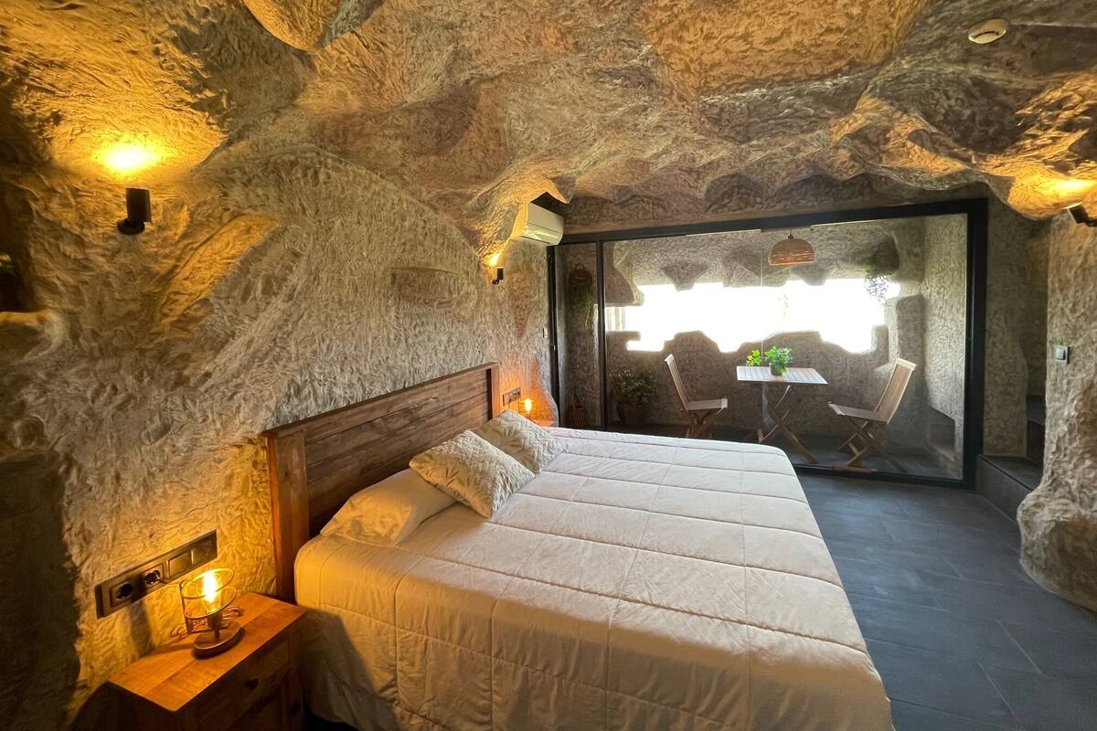 Een slaapkamer met een stenen muur, een houten bed, een nachtkastje en een tafel met stoelen.