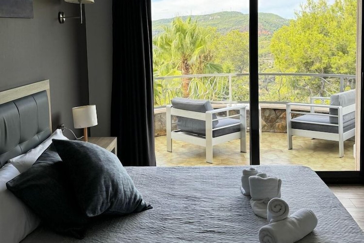 Un dormitorio con cama, dos lámparas de noche, vistas a un balcón con sillas y una mesa, y un cuadro en la pared.