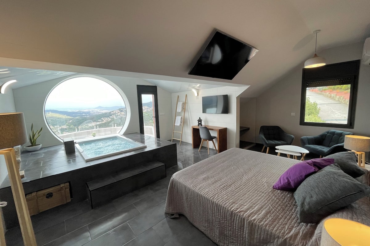 Een moderne slaapkamer met een groot bed, een bureau, een stoel en een klein tafeltje. Er is een televisie aan de muur bevestigd en een raam met uitzicht op de stad.