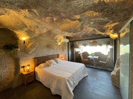 Quarto com parede de pedra, cama de madeira, mesa de cabeceira, mesa com cadeiras e vista através de uma porta de vidro.