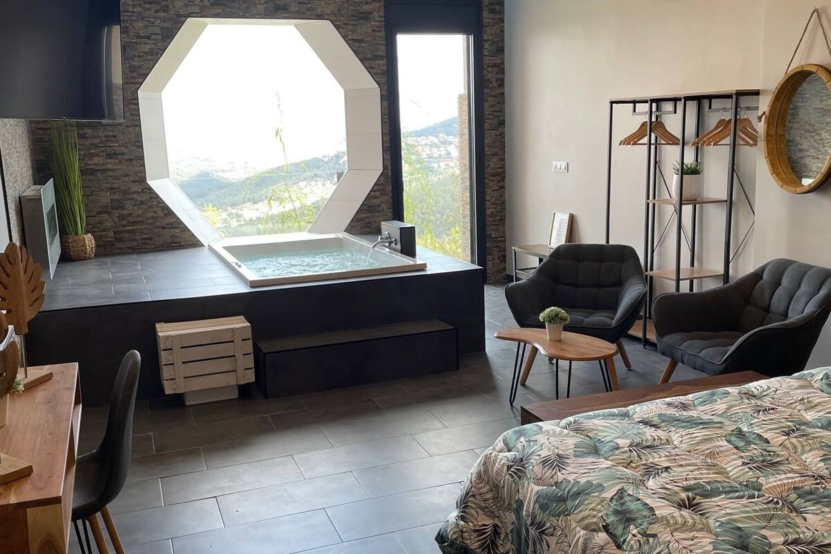 Een moderne slaapkamer met een groot bed, een zithoek met stoelen, een klein tafeltje, een groot raam met uitzicht en een inbouwbubbelbad.