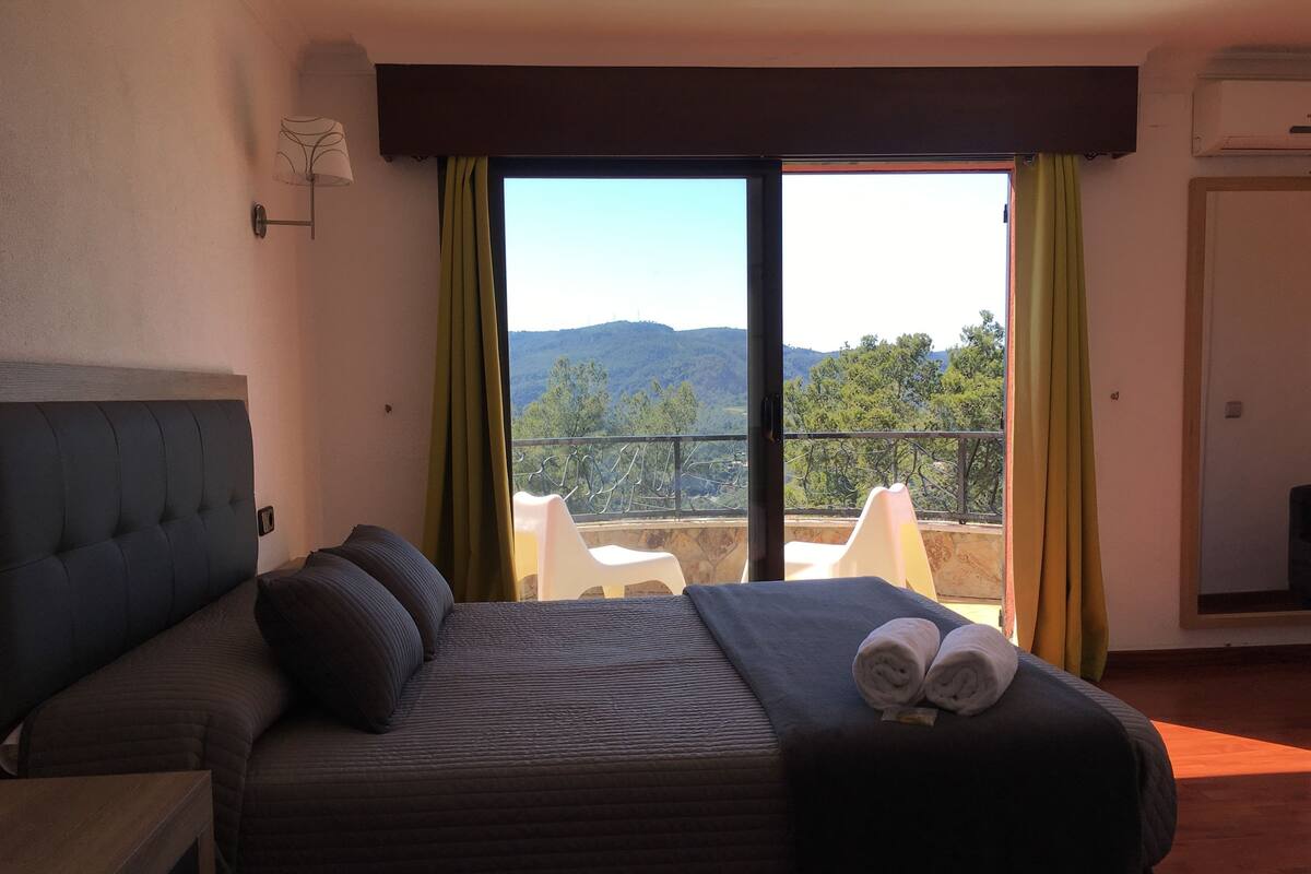 Een slaapkamer met een bed, nachtkastje, raam met uitzicht op de omgeving en een balkon met stoelen.