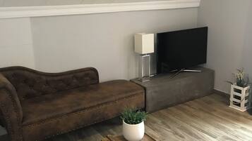 A loft-style living room with a brown leather sofa, a flat-screen TV mounted on the wall, a wooden coffee table with a plant, and a small white side table.