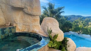A hot tub with a wooden deck, surrounded by large rocks and palm trees, overlooking a mountainous landscape. - El Petit Mirador (Torrelles de Llobregat)