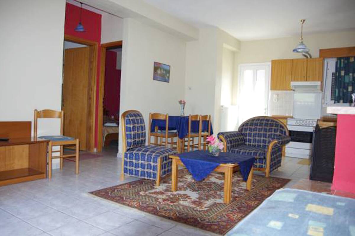 A living room with a sofa, chairs, a coffee table, and a rug. There is a kitchen area with a stove and cabinets.
