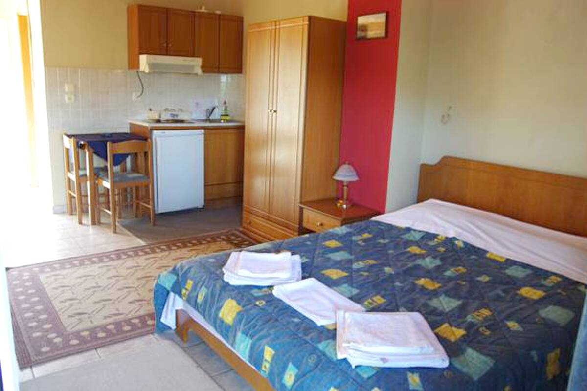 A compact hotel room with a bed, a desk, a kitchenette, and a red accent wall.