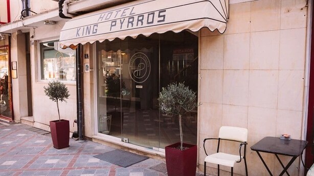 Un hotel llamado 'King Pyrrhos' con suelo de baldosas, dos plantas en macetas y una silla blanca en el exterior.