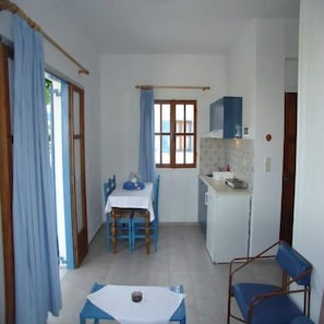 A compact kitchen with a dining area, a small refrigerator, and a window with curtains. - Hotel Agnanti (Milos)
