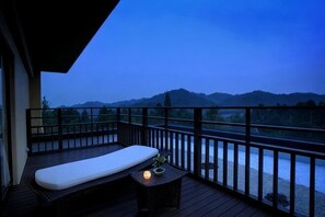 A balcony with a mattress, a small table with a candle, and a view of a river and hills.