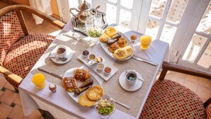 Une table de petit-déjeuner, dressée sur une terrasse offrant une vue sur l’extérieur, est garnie de divers aliments et boissons, notamment des viennoiseries, des fruits et des boissons diverses.