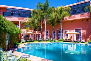 A pool area with lounge chairs and umbrellas, surrounded by palm trees and a building with arched windows. - Albakech Boutique Hotel & Spa (Marrakech)