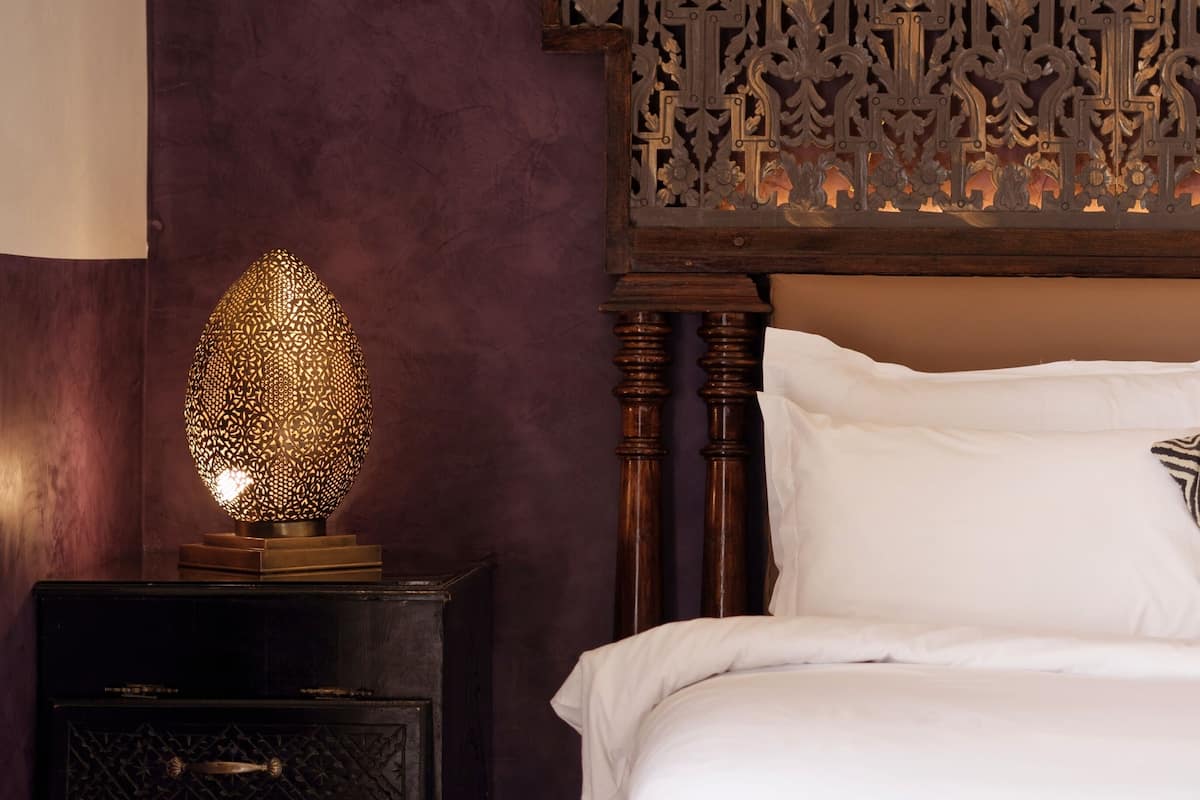 A bedroom with a dark wood headboard, a decorative wall panel, and a bedside table with a patterned lamp.