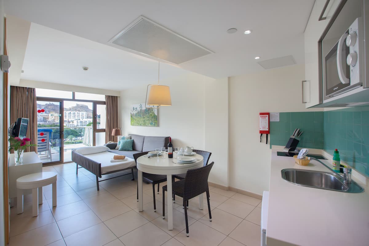 Quarto de hotel moderno com área de refeições, cozinha compacta e uma grande janela com vista para a cidade.