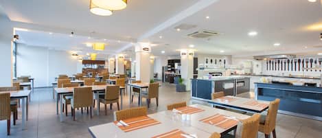 A modern dining area with multiple tables and chairs, a kitchen in the background, and overhead lighting.