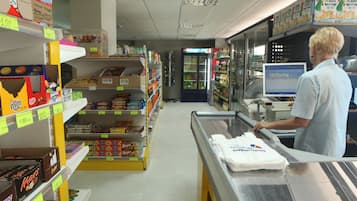 Interior de una tienda de comestibles con estantes llenos de productos variados, una vitrina refrigerada y una caja registradora con una computadora.