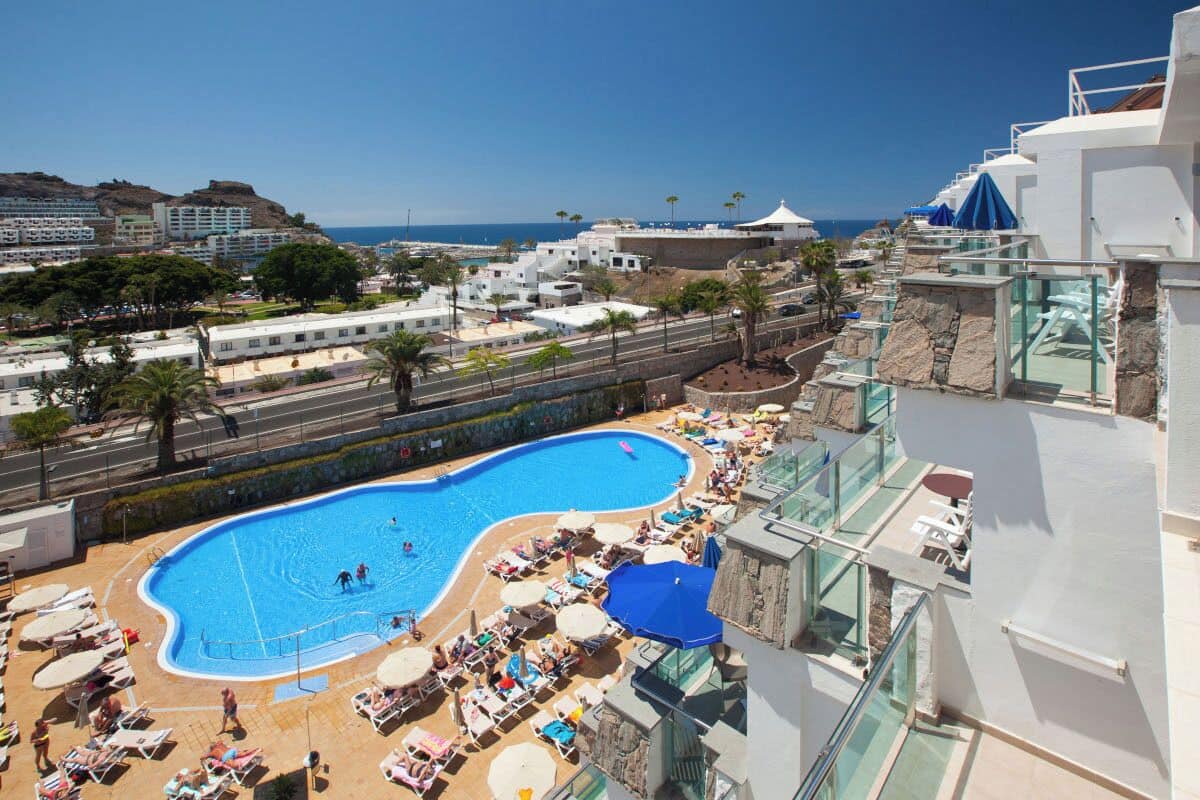 Um resort com uma piscina grande rodeada por espreguiçadeiras e guarda-sóis, vista desobstruída para o mar e montanhas ao fundo.
