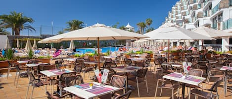 An outdoor dining area with tables and chairs, a pool in the background, and a building with balconies.