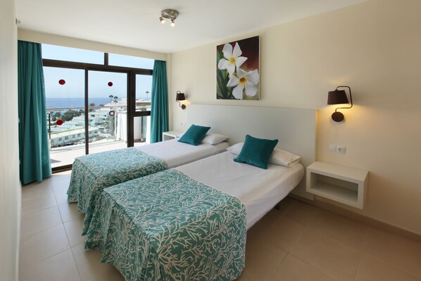 A hotel room with two beds, a large window with a view of the city, and a wall-mounted lamp. - Grupotel Revoli (Mogan)