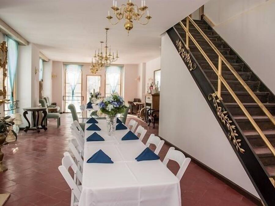 A formal dining room with a long table set for a meal, a staircase with golden railings, and a chandelier.