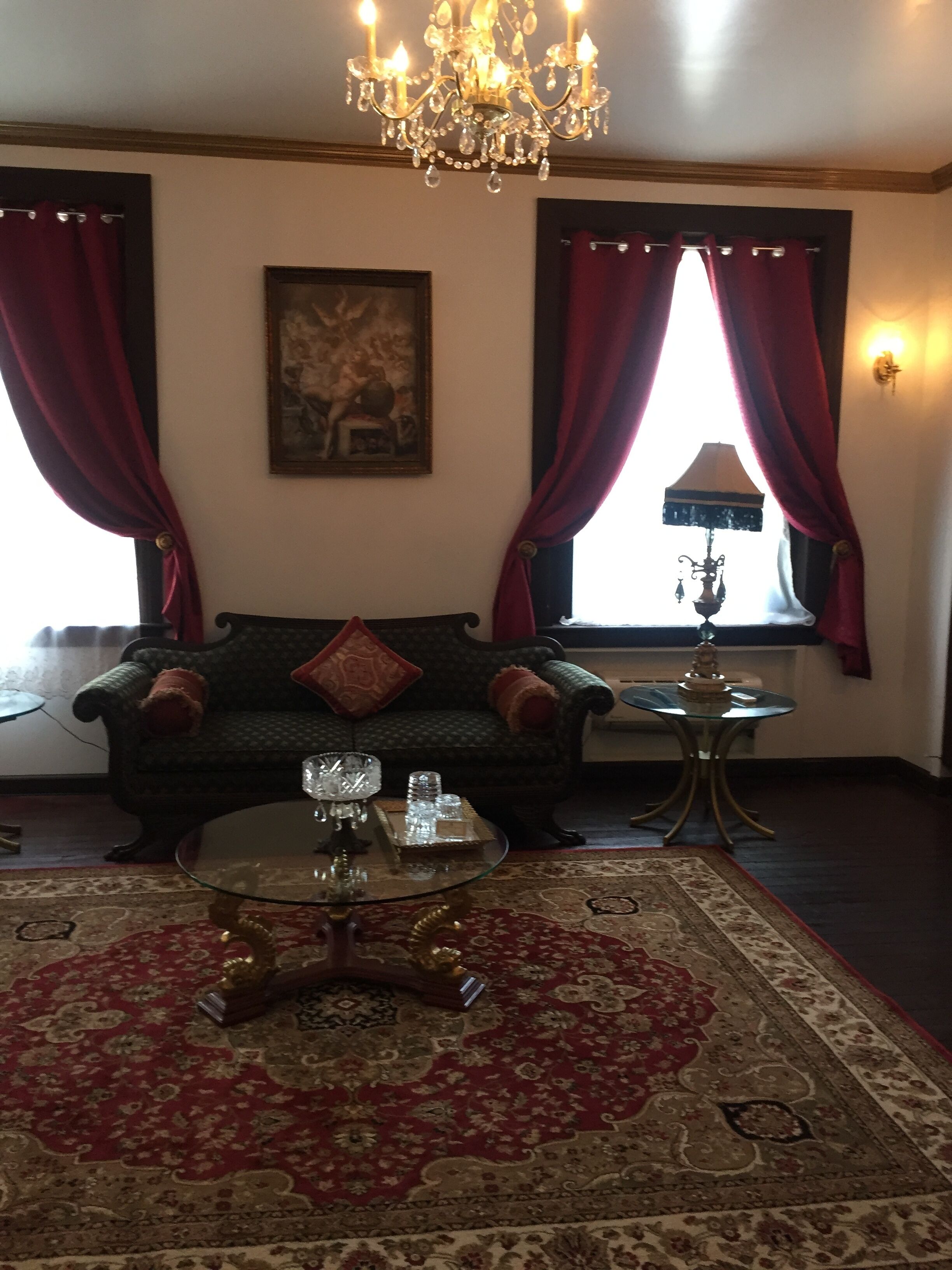 Een woonkamer met een groene bank, rode gordijnen, een kroonluchter, een glazen salontafel en een kamerbreed tapijt met een patroon.