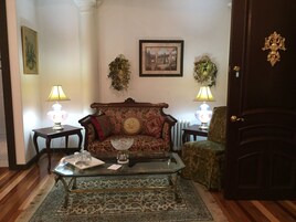 Una sala de estar con un sofá floral, una mesa de centro de vidrio, dos mesas auxiliares con lámparas, un cuadro enmarcado en la pared y una puerta de madera oscura.