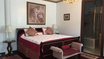 Une chambre avec un grand lit, deux tables de chevet, un lustre, un tableau et une porte-fenêtre coulissante donnant sur une salle de bain.