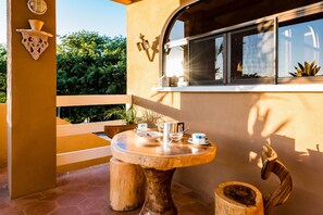 Una mesa redonda de madera con mantel blanco, rodeada de taburetes de madera, en un balcón con vista a vegetación.