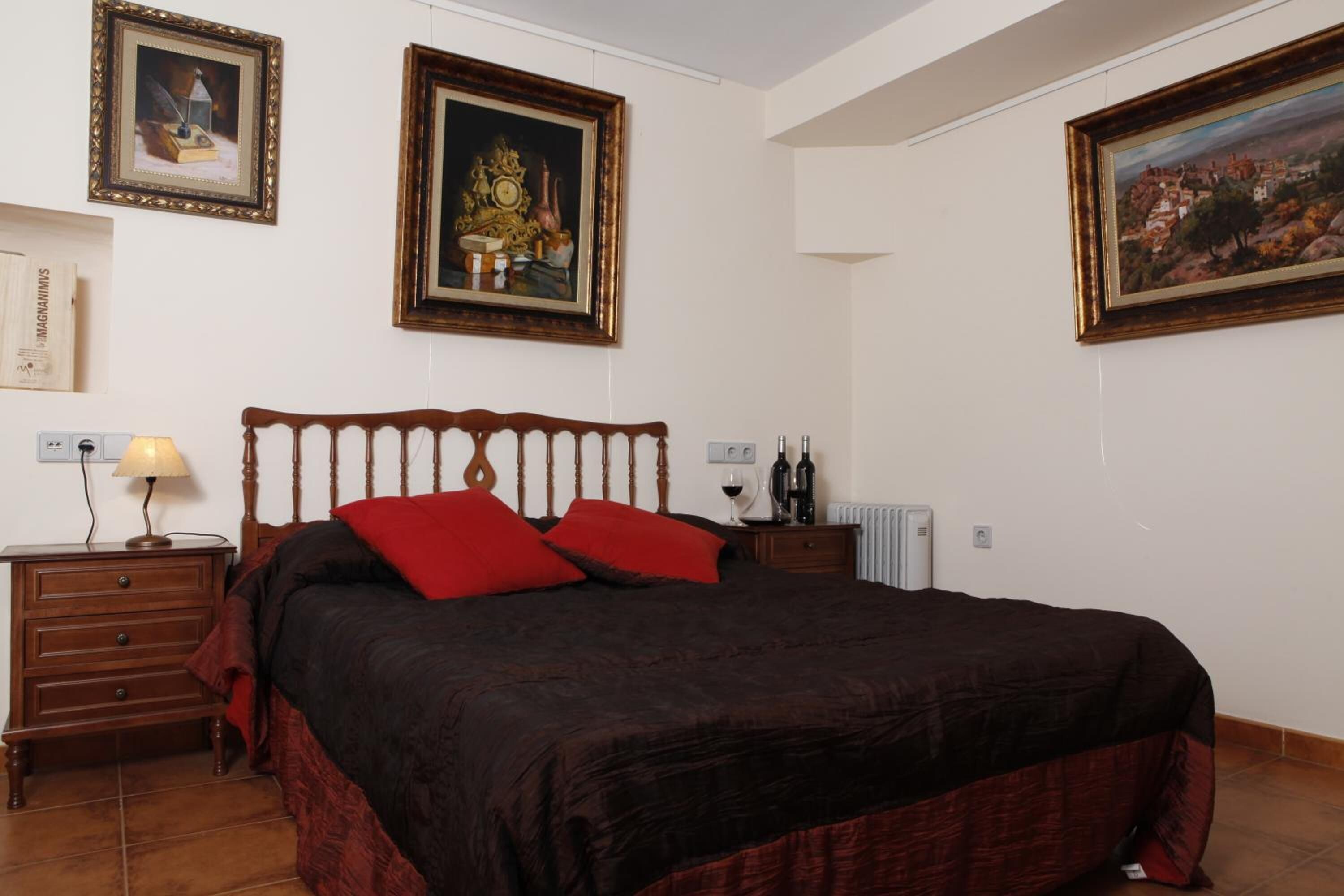 Ein Schlafzimmer mit einem hölzernen Bett, roten Kissen, einem Nachttisch mit Lampe sowie gerahmten Bildern an den Wänden.