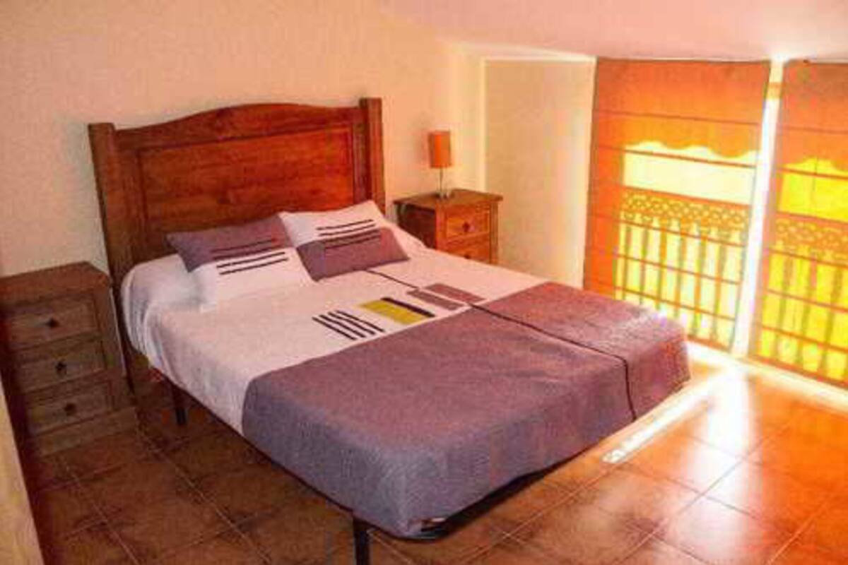 A neatly made bed with a wooden headboard, a bedside table with a lamp, and a view of a balcony with a railing.