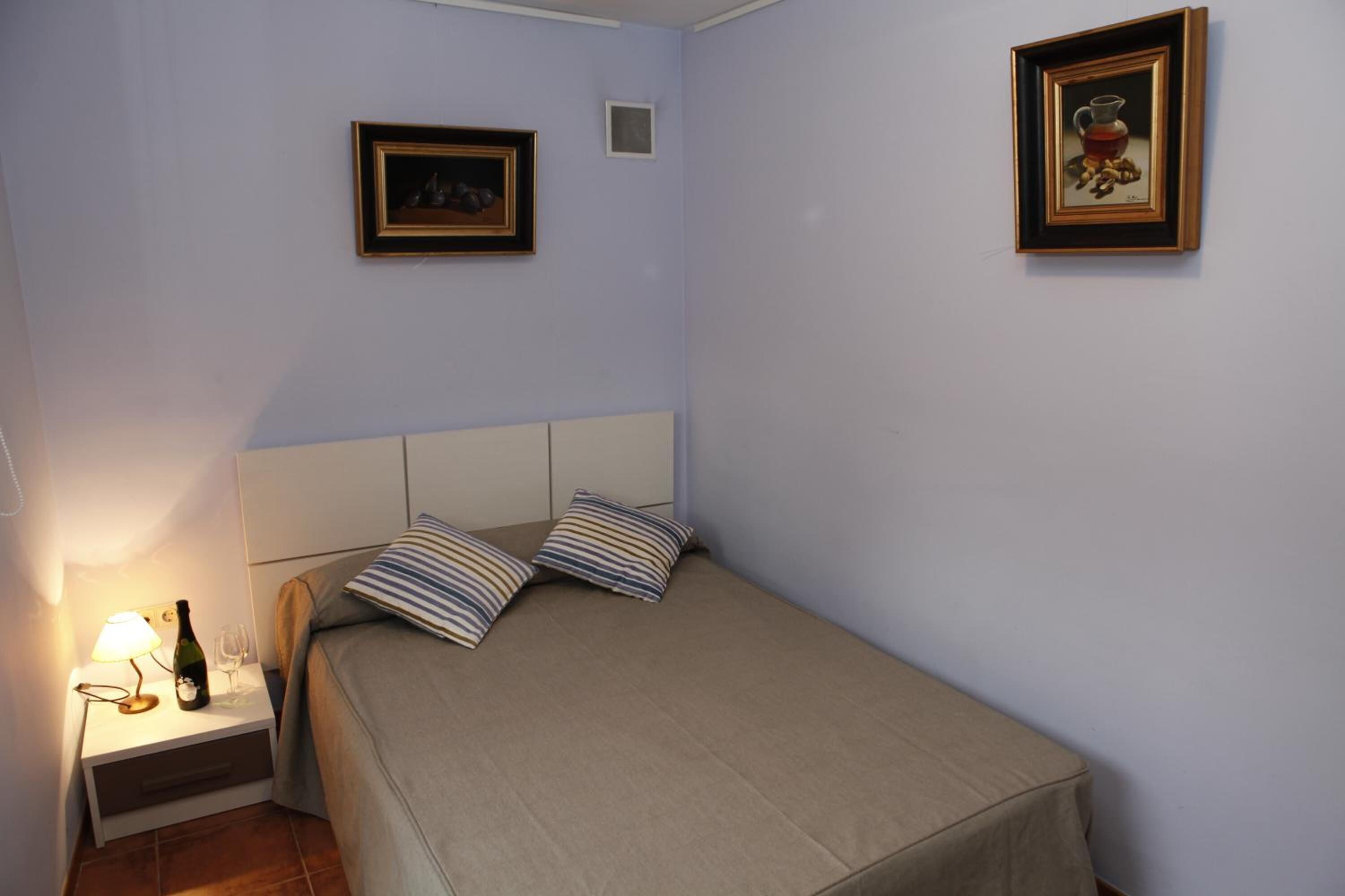 Un dormitorio con una cama, una mesita de noche, una lámpara, una botella y dos cuadros en la pared.