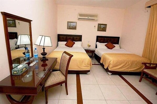 A hotel room with two beds, a desk with a mirror, a chair, and a bedside table with a lamp and a bottle. - Diamond 1 (Doha)