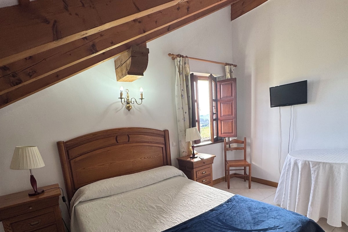 A bedroom with a wooden bed, a nightstand, a desk, a chair, a television, and a window with curtains.