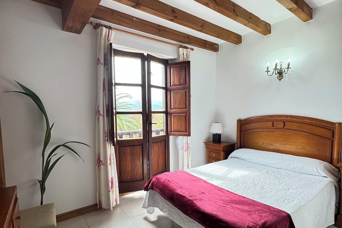 A bedroom with a wooden bed, a nightstand, a chair, a potted plant, and a window with curtains.