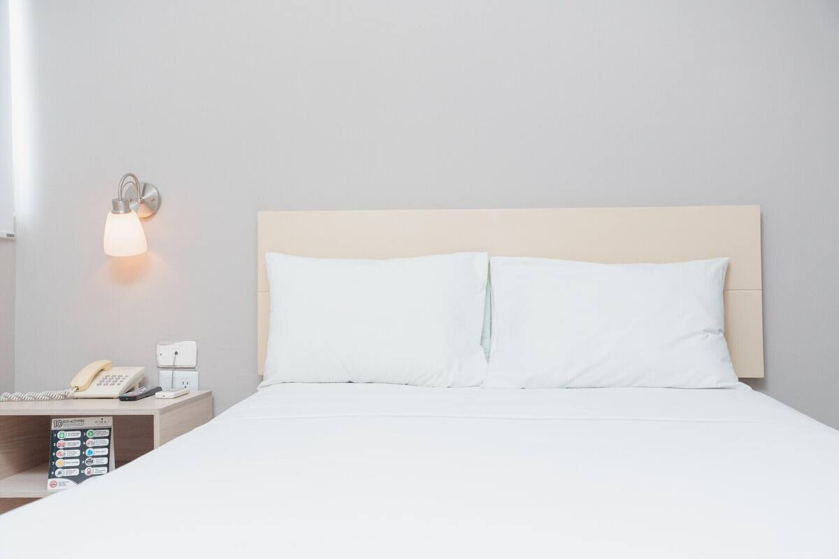 A hotel room with a bed, a nightstand with a phone and a lamp, and a wall-mounted light fixture.