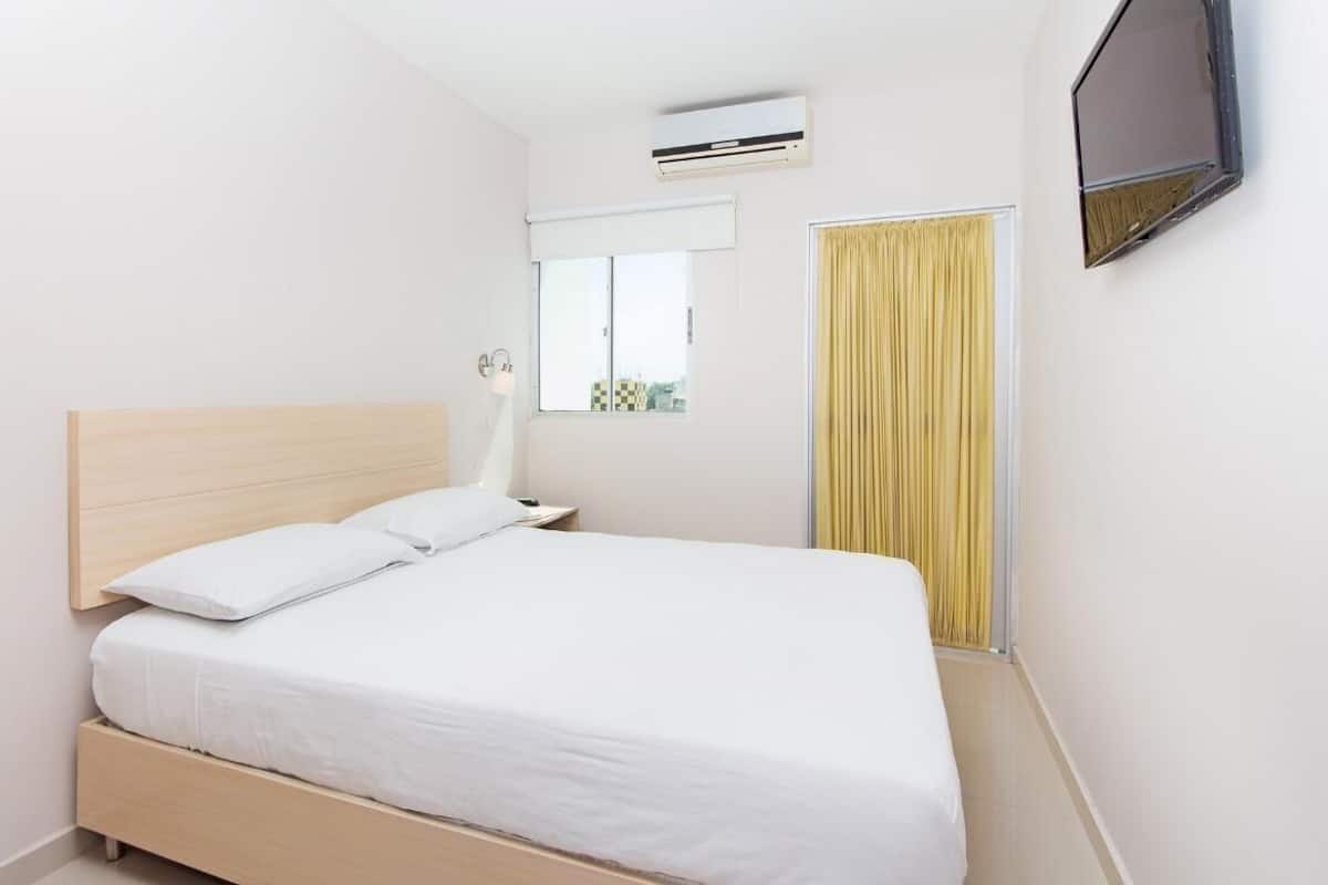 A hotel room with a single bed, a window with curtains, an air conditioning unit, and a flat-screen TV mounted on the wall.