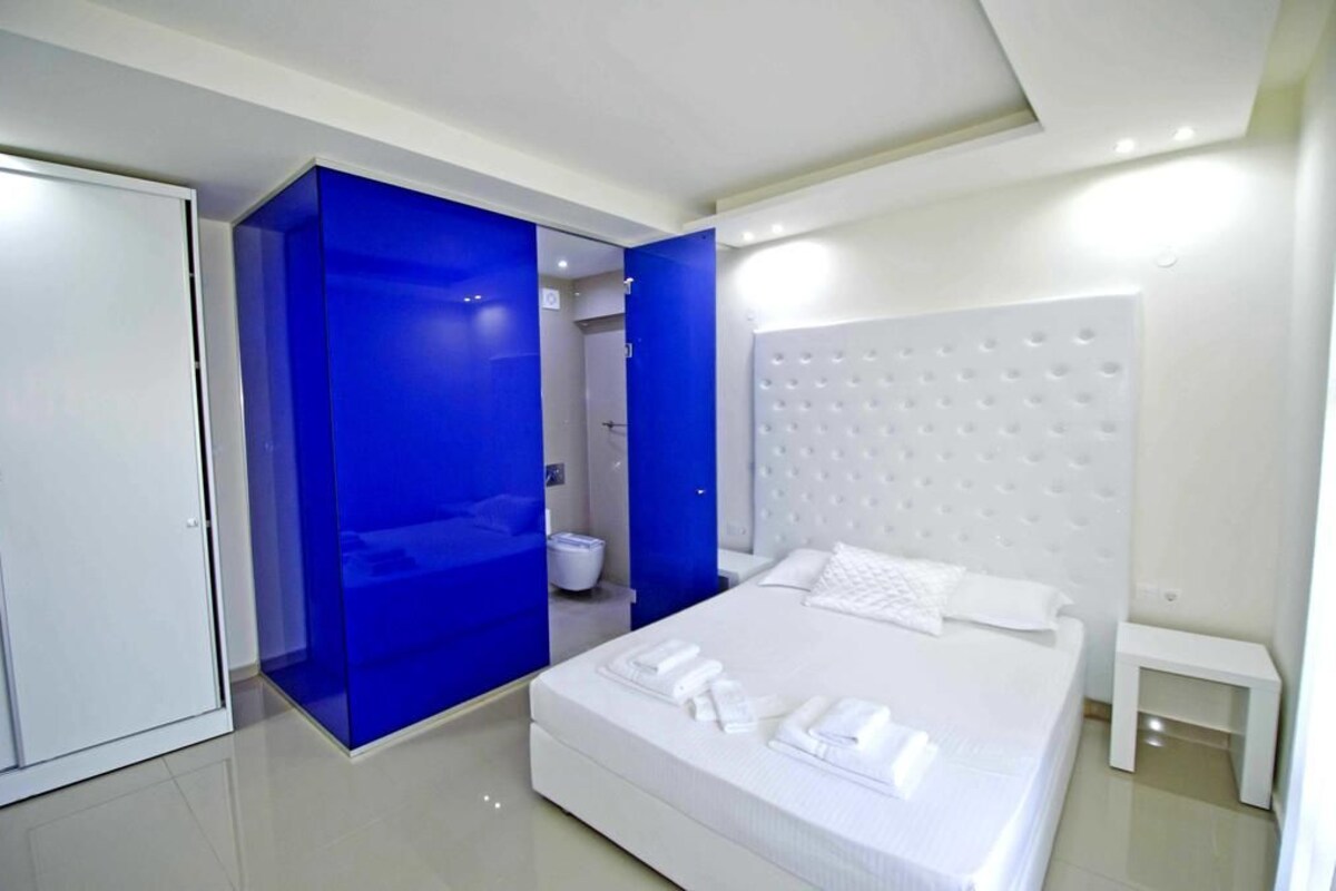 Quarto de hotel moderno com uma cama grande, cabeceira branca e uma parede de destaque azul.