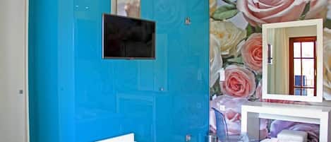 Una habitación con una pared de acento color turquesa, un televisor de pantalla plana montado sobre un banco blanco, un espejo con papel tapiz floral y una cama con toallas dobladas con cuidado.